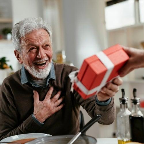 A bearded man sitting at a table accepts a wrapped gift with a big smile
