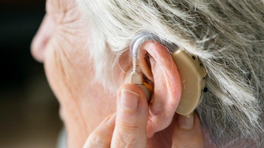 White older adult woman touches a hearing aid in her left ear