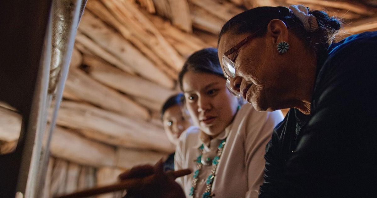 A senior Native American woman is showing a younger generation how to weave.