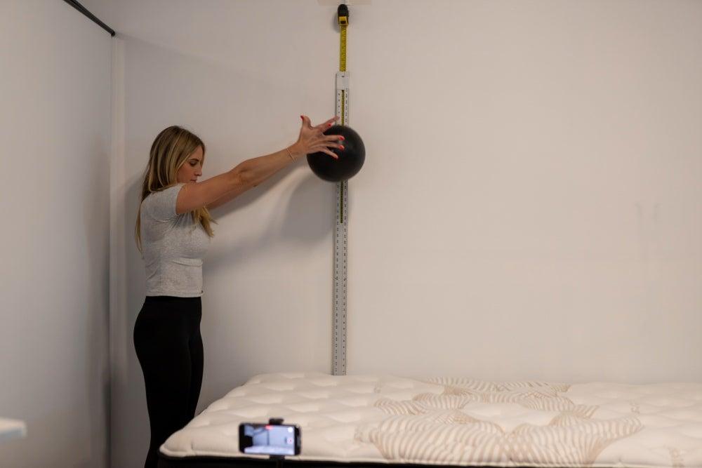 A woman holding a weighted ball against a measuring tape, about to drop it on a bed