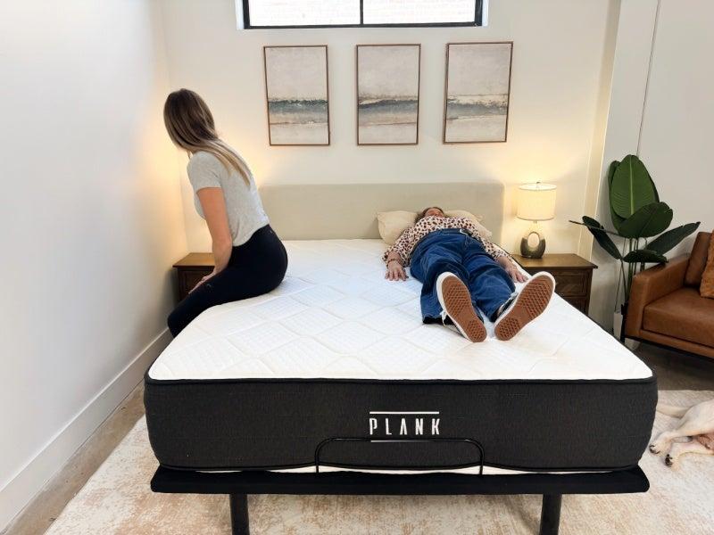 A woman sits on the edge of the Plank Firm Luxe mattress while a tester lies on the other side