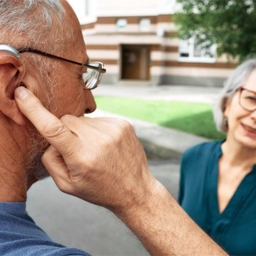 A man is talking to a woman outside with a finger on the hearing aid in his ear.