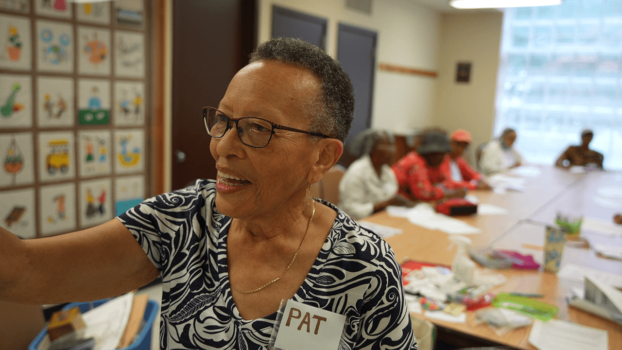 An older woman named Pat, wearing glasses and a patterned blouse, stands at the forefront of a classroom setting with a smile. Behind her, several other older adults are seated at a long table, engaged in an activity involving colorful materials. The room has bright lighting and educational posters on the walls.