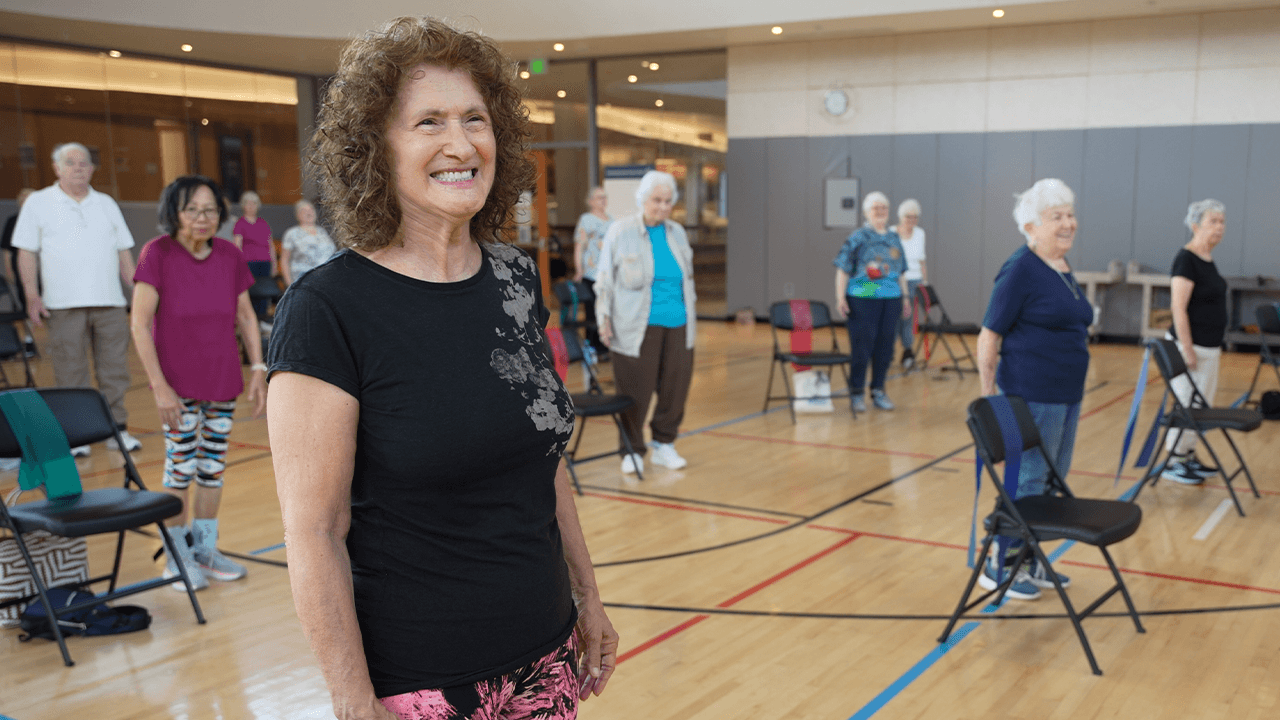 A group of older adults is participating in a fitness class in a gymnasium. They are standing and smiling, with chairs spaced out around them for support. The foreground features a woman with curly hair wearing a black t-shirt and pink patterned pants, smiling at the camera. The gym has a wooden floor and large windows in the background.