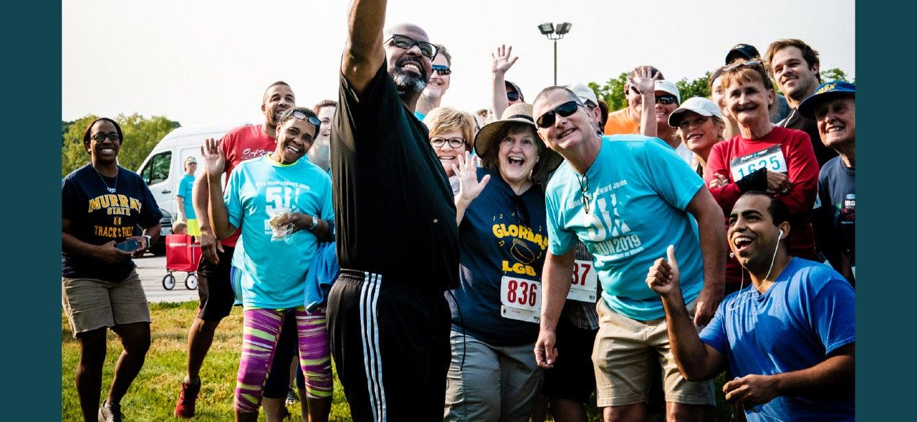 A diverse group of people, including both older and younger adults, pose together outdoors at a community event or race. Many are wearing numbered race bibs and colorful athletic clothing. A man in the center, wearing black, raises his arm in celebration as others around him smile and cheer. The group exudes energy and enthusiasm, suggesting a positive and inclusive community gathering.