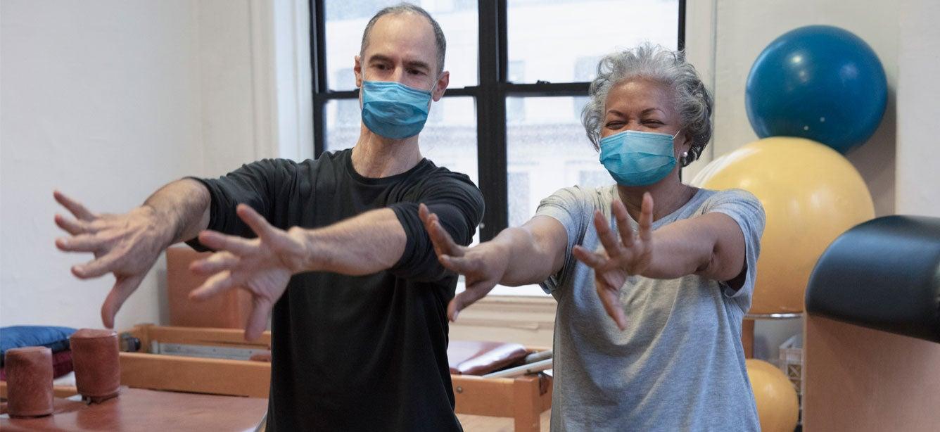 A senior Black woman is doing stretching exercises with her instructor in a gym.