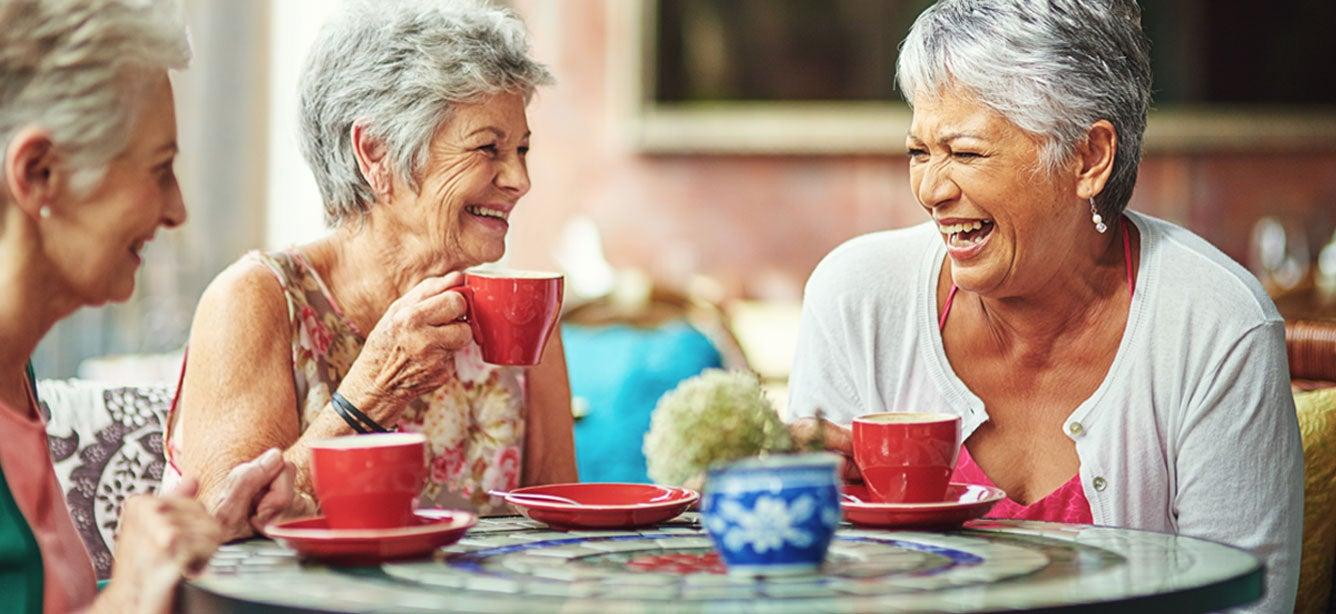 Three older women laughing and enjoying coffee at a colorful table with red cups and plates in a relaxed setting.