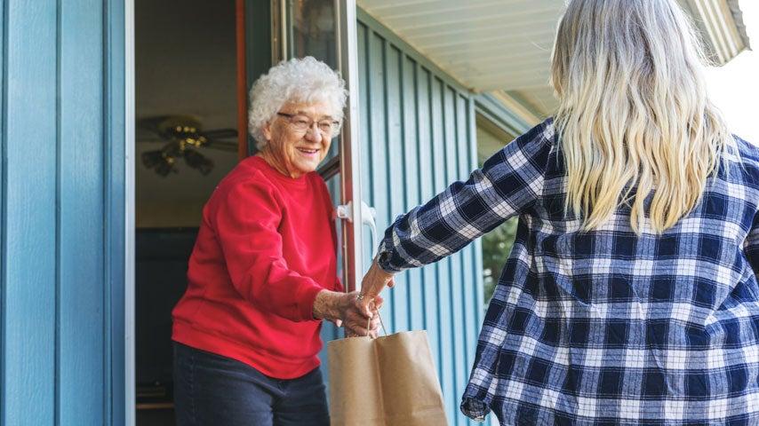 A happy senior woman wearing glasses and a red sweater is receiving a bag of groceries at her door from an Uber Eats driver.