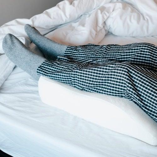 A person's legs in checked blue pajamas rest on a knee pillow in bed