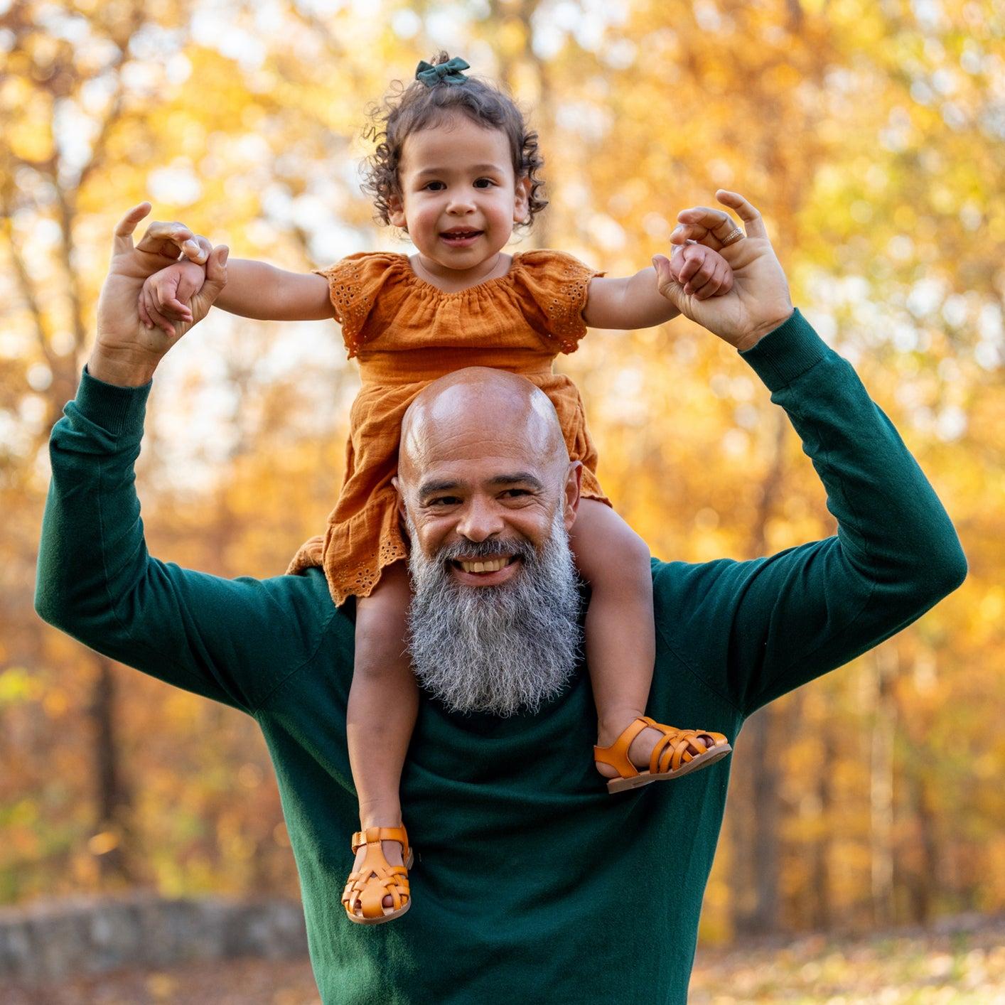A black older man with a graying beard is holding his grandchild on his shoulders and both are smiling on a fall day.