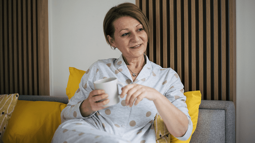 smiling older woman in pajamas sits on couch holding coffee cup