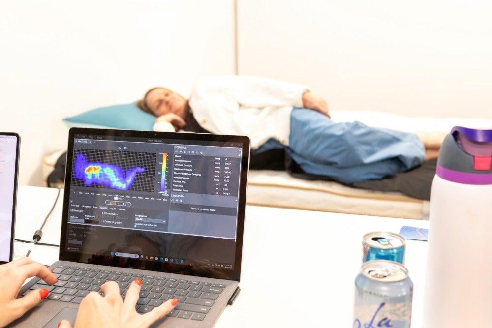 Woman lies on her side on a bed in the background while in the foreground a computer measures pressure buildup on the mattress