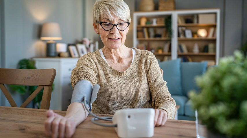 older woman seated at wood table checking blood pressure with home monitor