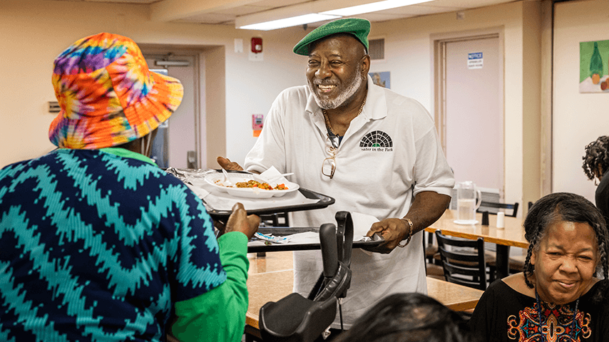 smiling older man with Center in the Park shirt handing meal tray to another older adult