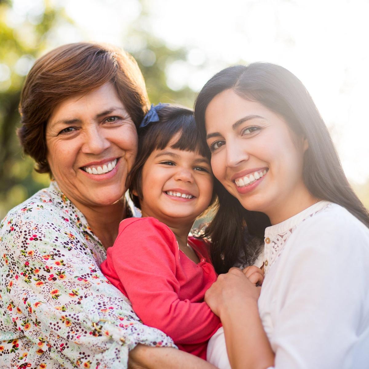 Three generations of women smiling outdoors: a grandmother, mother, and young girl embracing each other