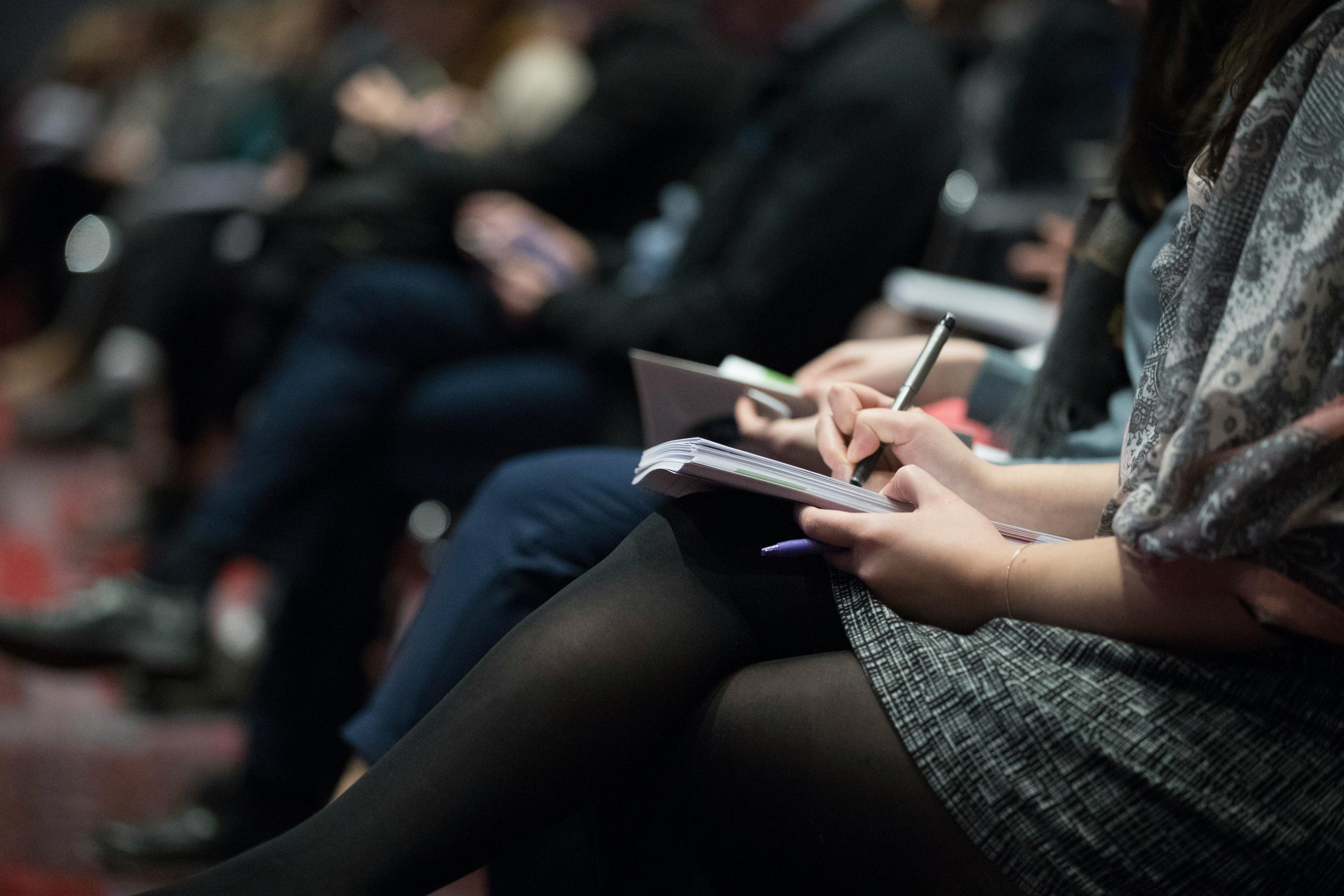 People taking notes during a conference session, focusing on a speaker's presentation.