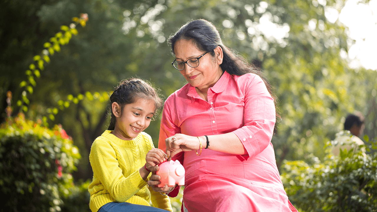 A woman wearing glasses and a bright pink kurta is sitting outdoors with a young girl in a yellow sweater. They are both smiling and focused on a pink piggy bank, which they are holding together. The background is a lush, green park with trees and shrubs, giving a peaceful and pleasant atmosphere.
