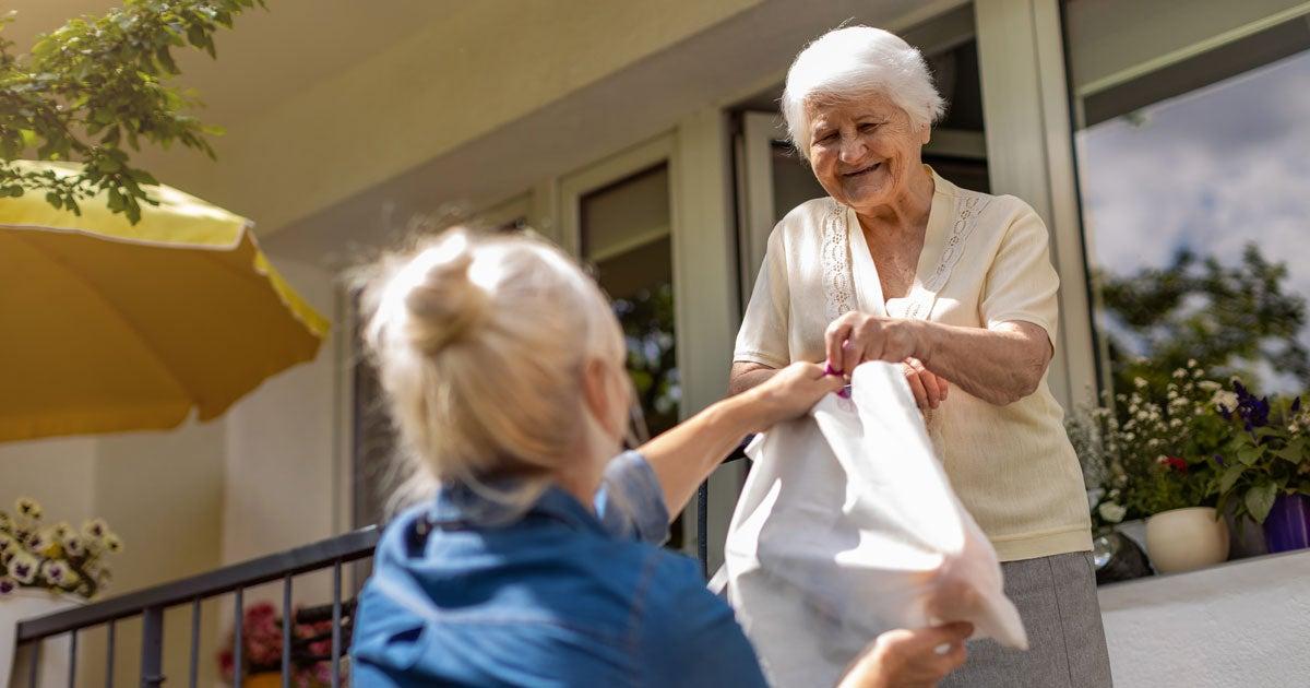 A middle-aged female Caucasian caregiver is handing groceries to her older mother while she's on the porch.
