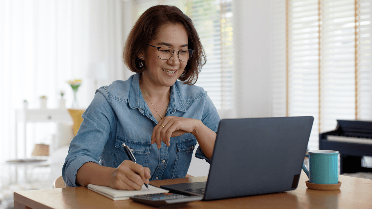 An older woman with glasses, wearing a denim shirt, smiles while working on a laptop at a wooden table. She is taking notes in a notebook and has a calculator and a blue mug on the table. The background features a bright, well-lit room with a piano and some home decor items.
