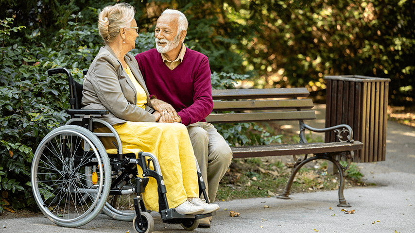 older white couple in park, woman seated in wheelchair and man on bench