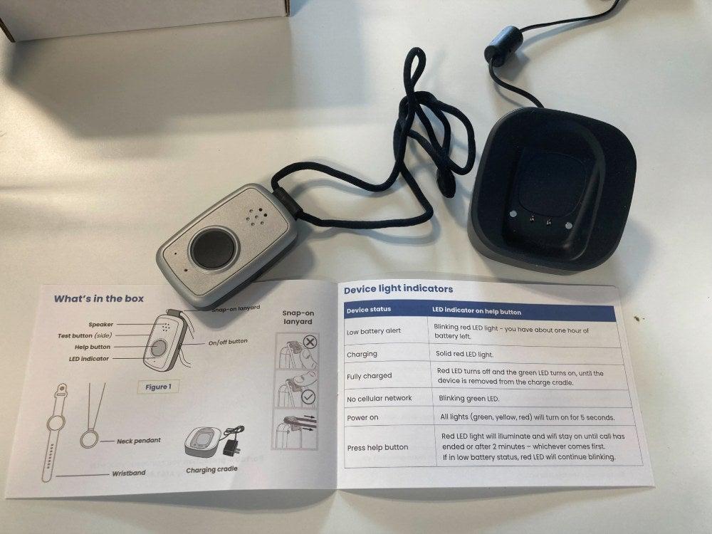 The MobileHelp Solo device with a lanyard attached, its charging cradle, and an open user manual showing what’s in the box and the meaning of light indicators