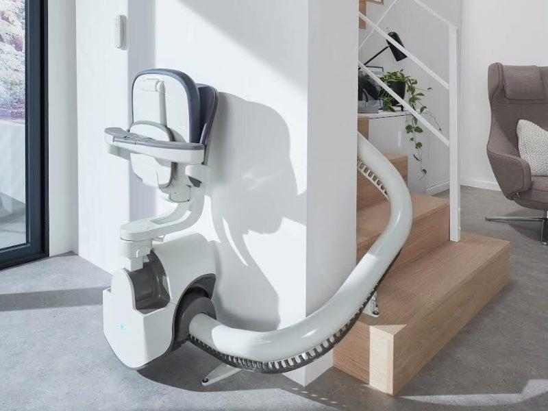 An Access BDD stair lift, with the seat folded up, sitting at the bottom or the stairs in a modern room