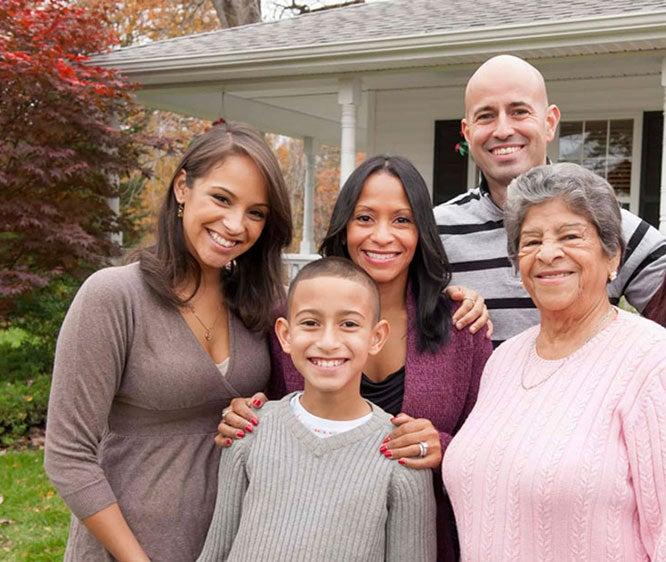 A multigenerational family stands together outdoors in front of a white house. The group includes an older woman with gray hair, a middle-aged man with a bald head, two women with dark hair, and a young boy in the front. They are all smiling and appear happy, with autumnal trees and a porch visible in the background.