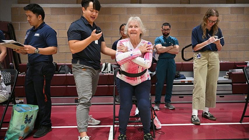 Woman practices a sit-to-stand exercise with her hands crossed across her chest, squatting down into a chair with the assistance of a professional.