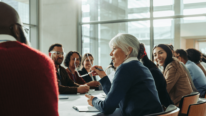 adults gathered around conference table listening to older woman speak