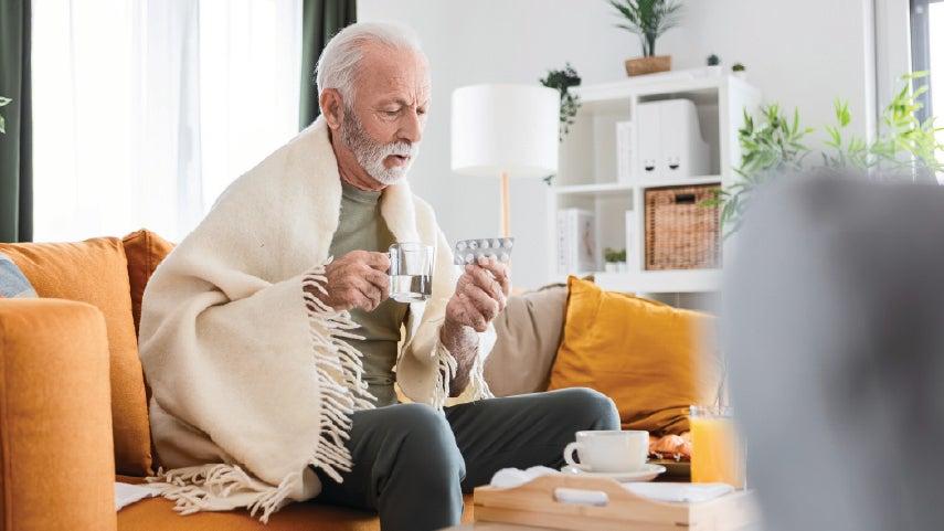 A white senior man is holding his hot tea and medicine in his hand while trying to sit on a couch with this blanket. He's sick with RSV.