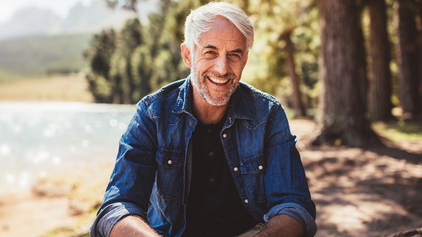 smiling man with gray hair and denim shirt sitting outside next to lake