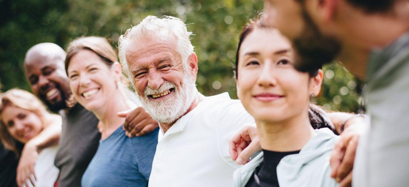 A diverse group of six smiling people standing shoulder to shoulder outdoors, with greenery in the background.
