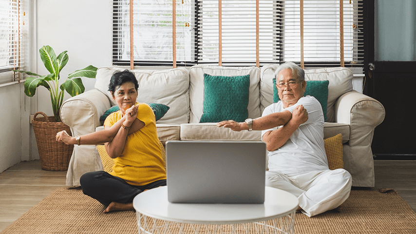 Older man and woman stretching on floor while looking at laptop propped in front of them