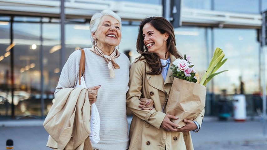 two women smiling women arm in arm, one with short gray hair, the other with long brown hair carrying a grocery bag