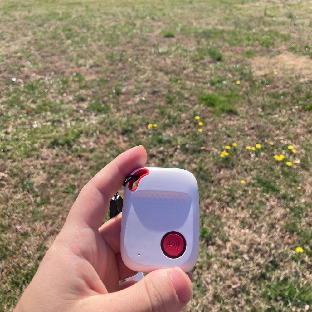 A hand holding a white Bay Alarm SOS All-in-One in a grassy field
