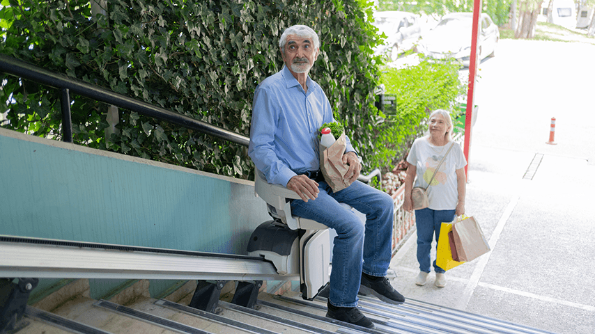 Man on outdoor stair lift holding a bag of groceries as woman with shopping bags watches him from bottom of stairs
