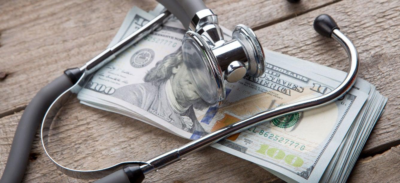 A doctor's stethoscope sits on top of a stack of one hundred dollar bills.