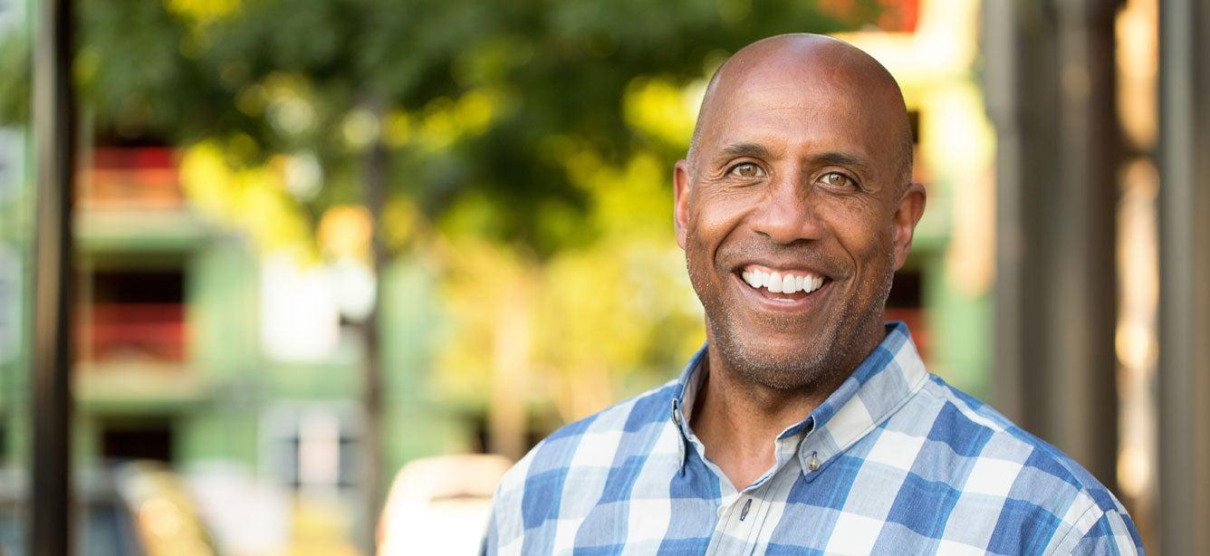 Smiling man in a blue and white checkered shirt stands outdoors with green trees and blurred buildings in the background.