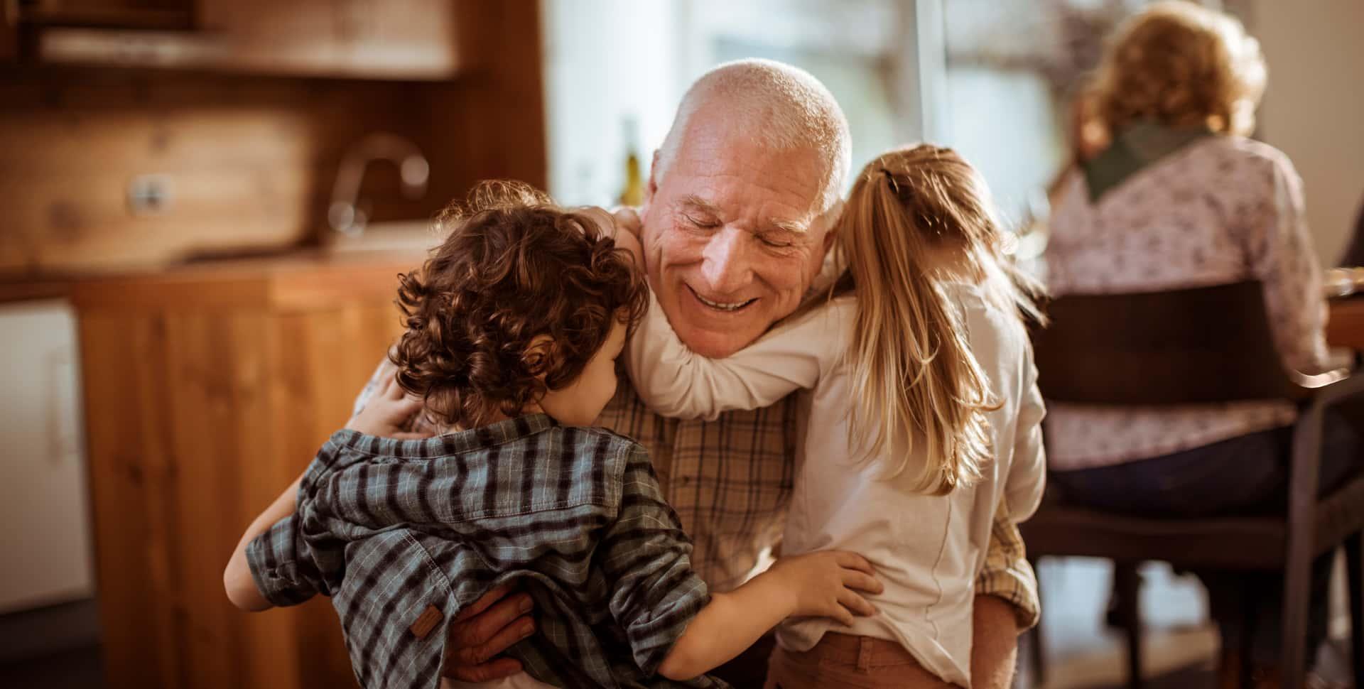 A grandfather is hugging his two grandchildren in the kitchen of his daughter's home.