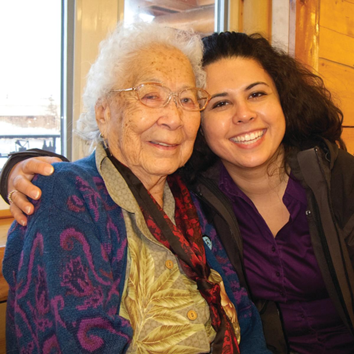 A senior Hispanic woman is hugging her granddaughter, both are smiling.