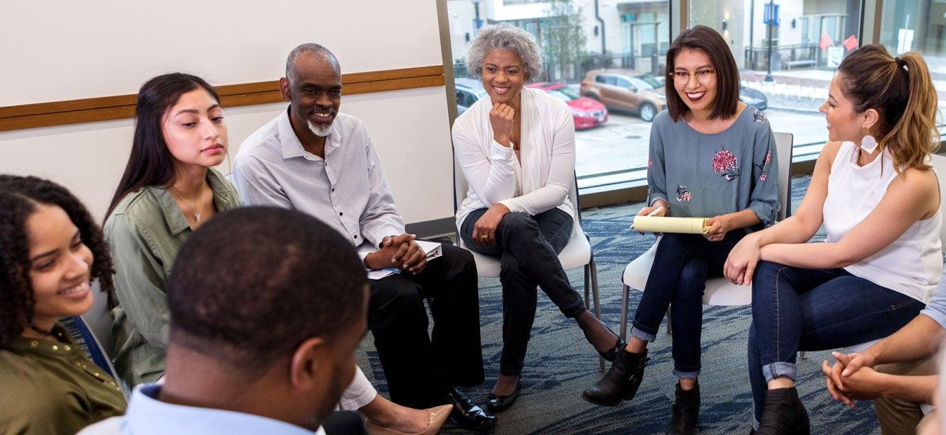Group of diverse adults seated in a circle, engaged in conversation, smiling, with a notepad in hand.