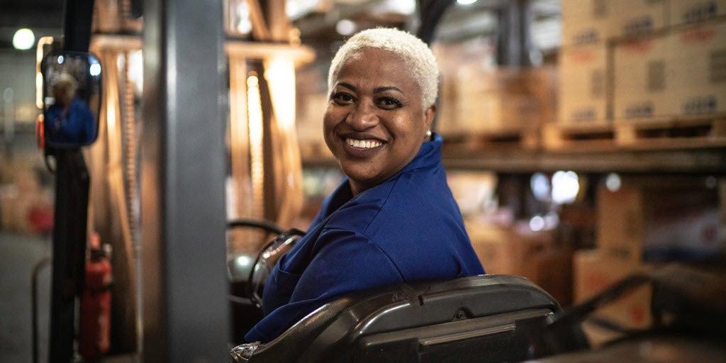 An older, black female is smiling while on the job in a production warehouse.