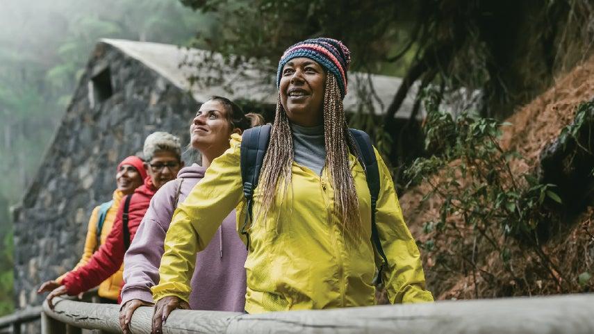A group of older women are on a hike in the outdoors, the person is front is a Black women wearing a yellow coat.
