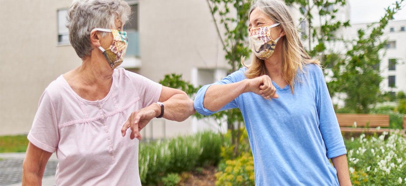 Two older women, standing outside and visiting during the pandemic, are elbow bumping while wearing masks.