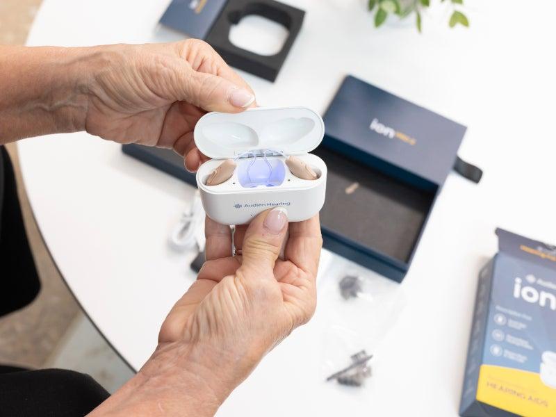 Hands hold the Audien Ion Pro 2 hearing aids in their charging case