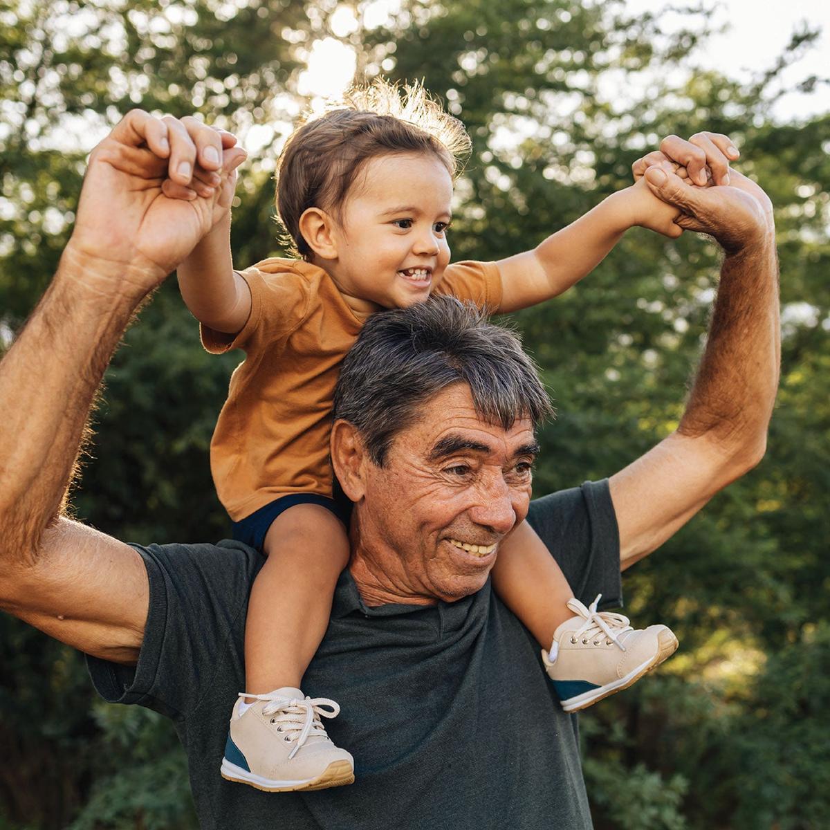 Hispanic grandfather is outside holding his grandson on his shoulders. They're both smiling.