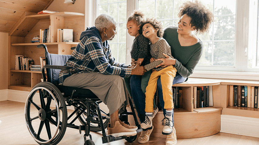 grandmother in wheelchair holding smiling grandchildren's hands while their mom hold the children on her lap