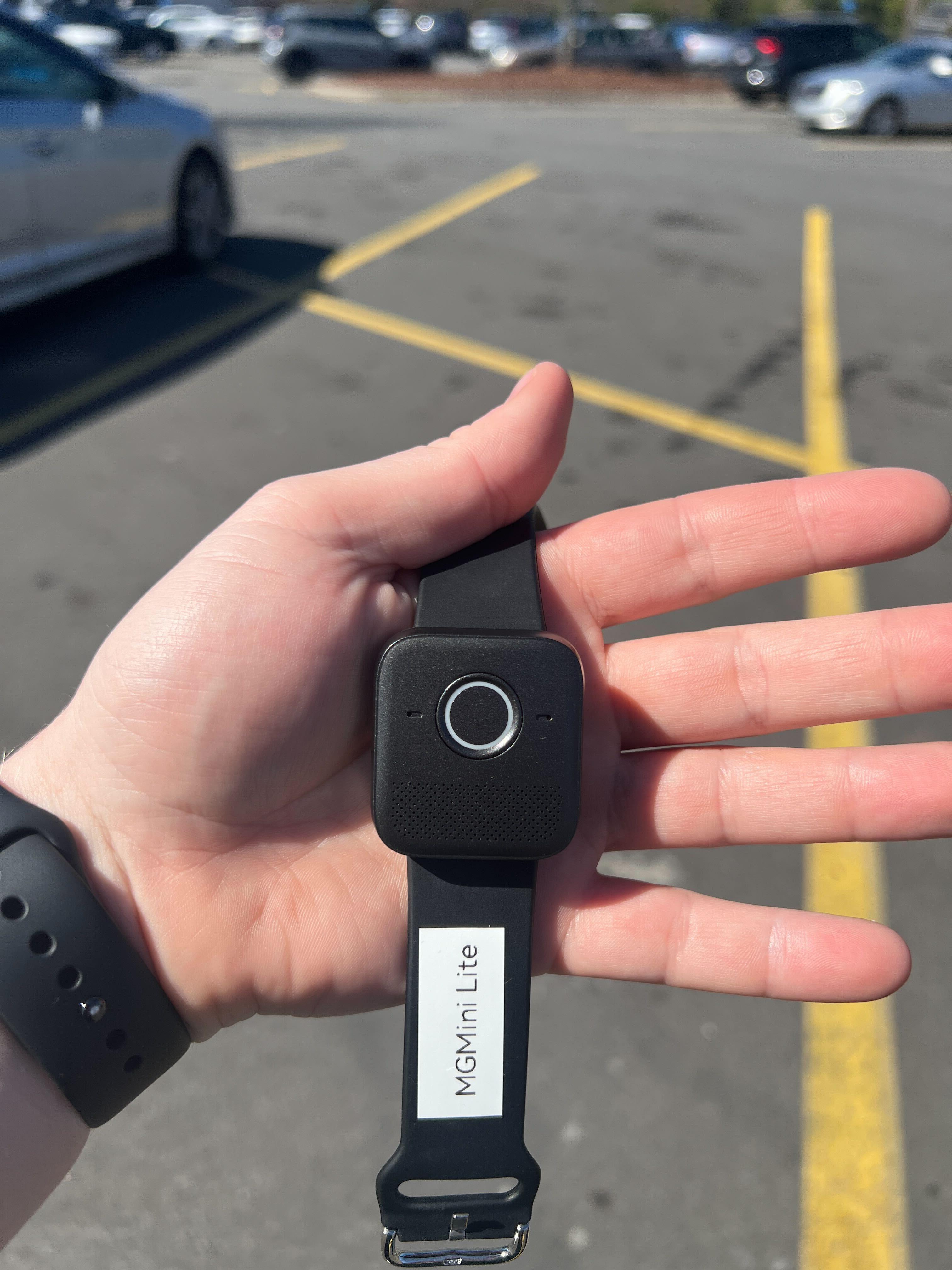 A man holds a small black medical alert watch in his left hand in a big-box store parking lot