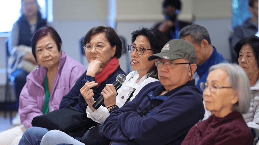 Older adults seated in an audience as speaks into handheld microphone