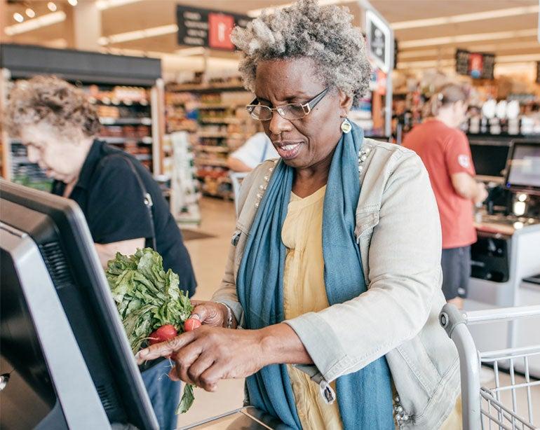 A Black senior woman is seen checking herself out at the grocery store, processing radishes over the payment kiosk.